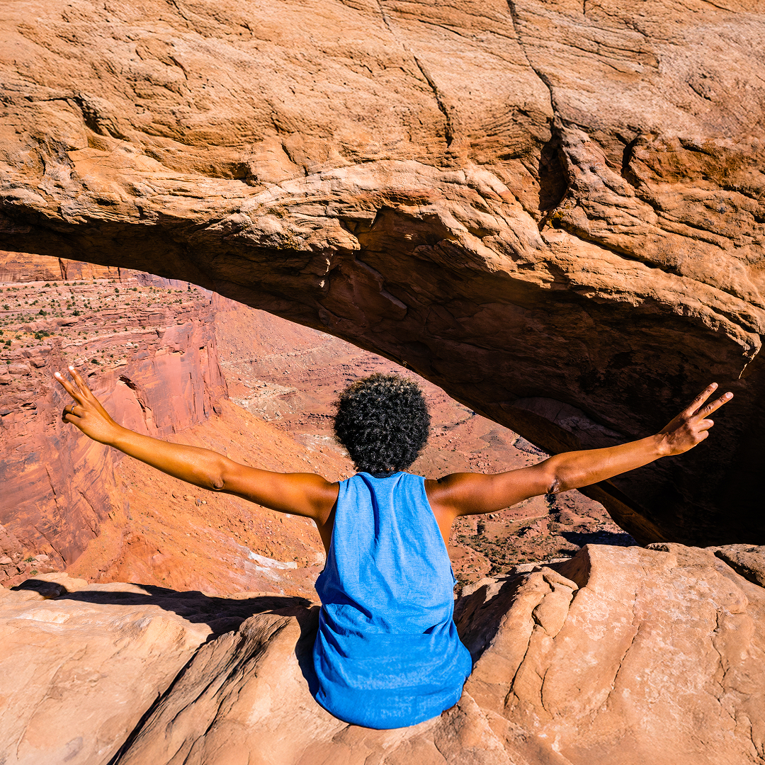 Person sitting on red rock ledge under a natural arch in Utah, raising arms in celebration during a Grand Circle road trip. Person sitting on red rock ledge under a natural arch in Utah, raising arms in celebration during a Grand Circle road trip.
