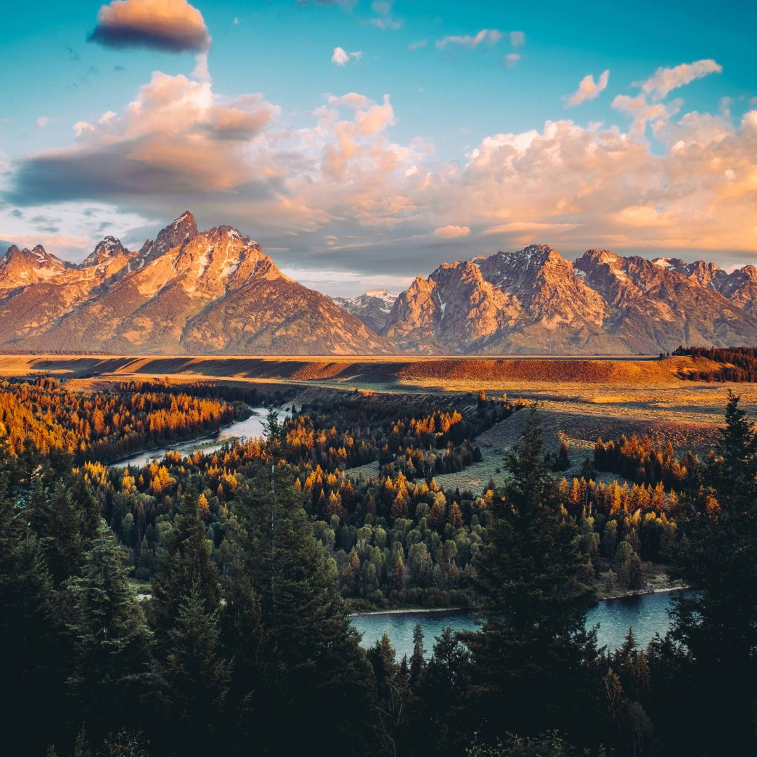 Grand Tetons to Yellowstone road trip view of sunlit peaks over a winding river and autumn forest in Wyoming