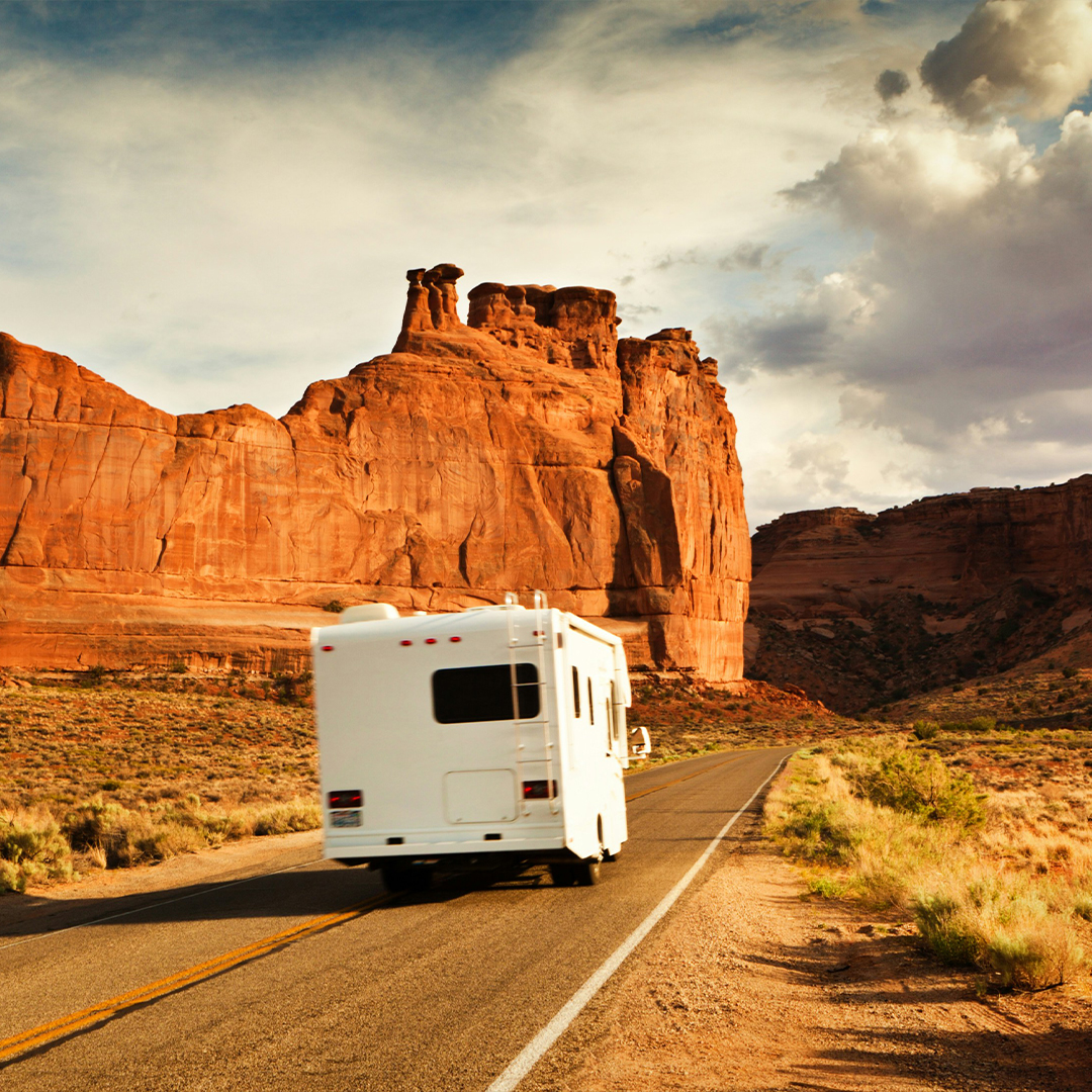 An RV on the road winds through desert cliffs and red rock formations at sunset in the American Southwest.